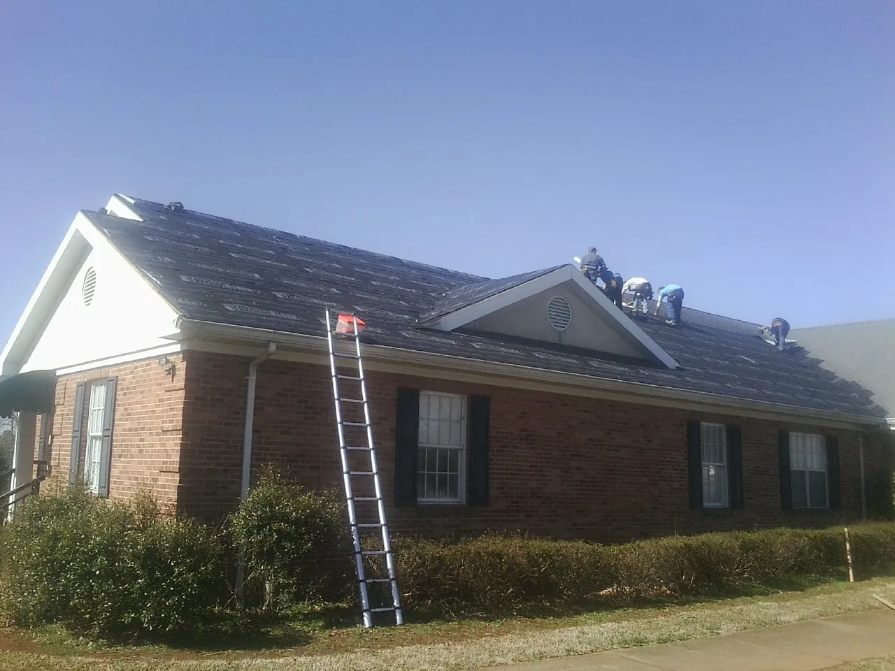 Crew tearing shingles off a brick ranch roof with a ladder set — Metal Roof Repair work by our crew in Pineville