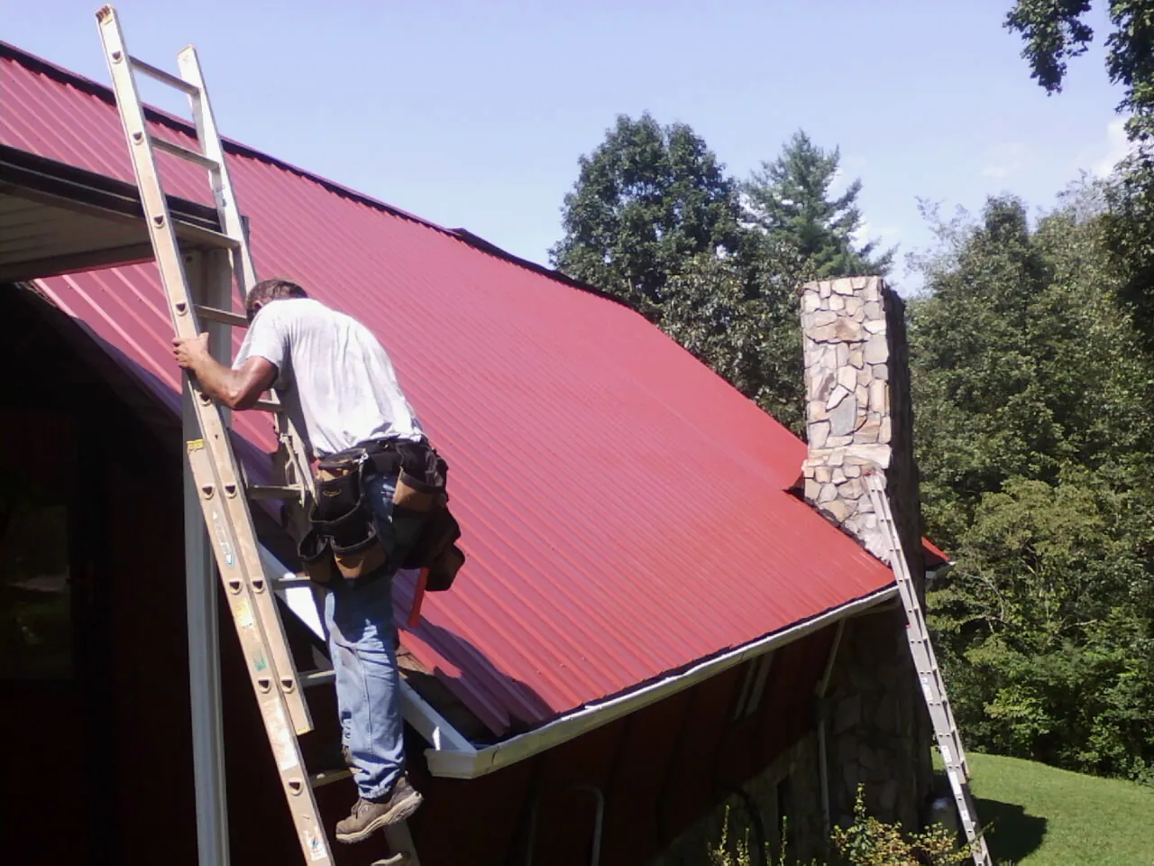 Roofer on a ladder beside a red metal roof and stone chimney — Soffit & Fascia Repair by Roofing Brothers in Pineville
